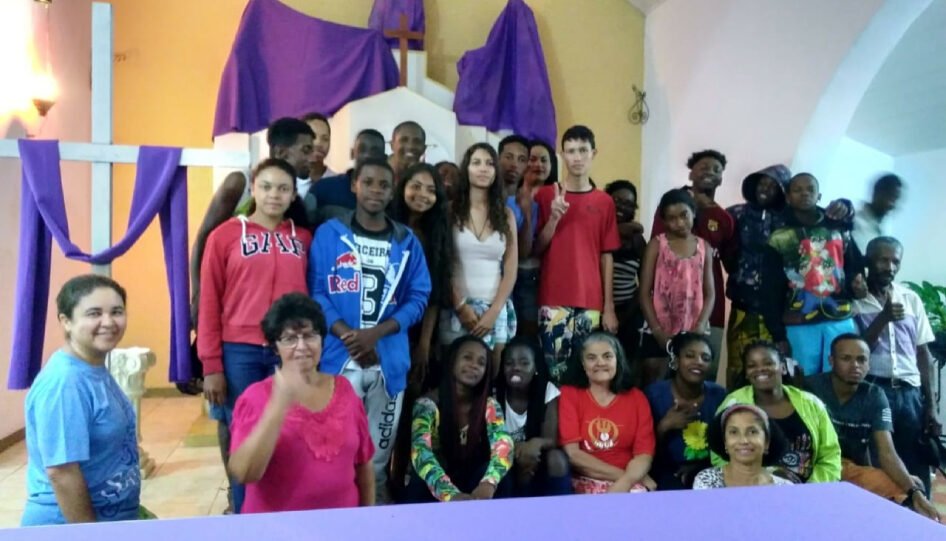 Grupo grande de pessoas em uma igreja. Algumas estão ajoelhadas e outras estão de pé.