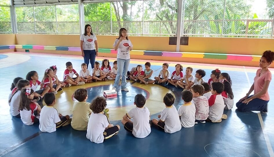 Uma mulher fala para um grupo de crianças, que estão sentadas em roda, fazendo um círculo em torno dela.