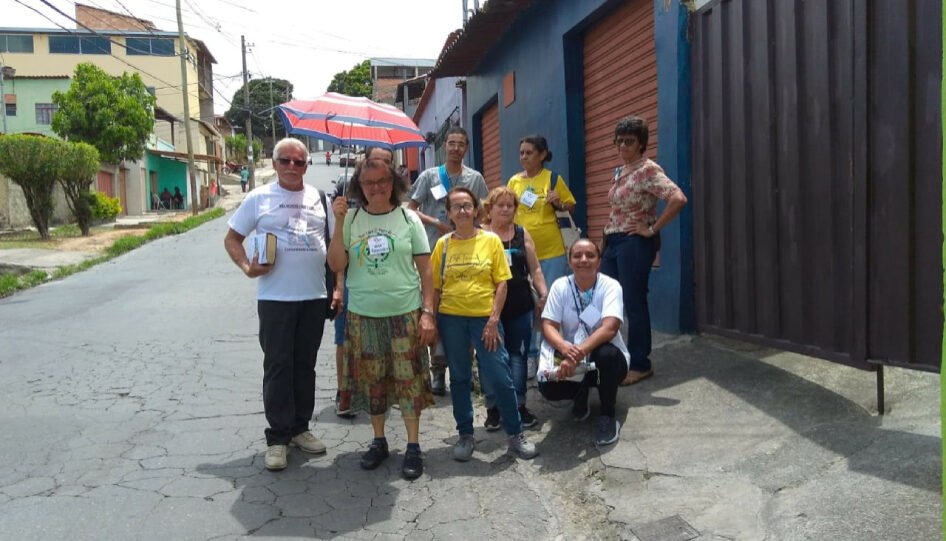 Grupo de nove pessoas, parado em frente a uma casa. A mulher da frente segura um guarda sol.